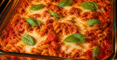 Baked rotini pasta casserole with melted mozzarella cheese and fresh basil in a glass dish inside an oven.