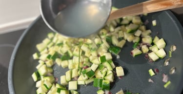 A metal ladle pouring broth over diced zucchini and onions cooking in a skillet.