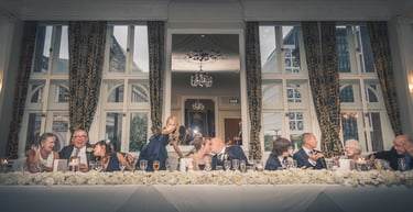 Wedding party at a floral head table with a kissing bride and groom in an elegant ballroom.