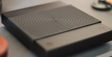A black Acaia Lunar digital coffee scale with a textured heat resistant mat on a grey surface.