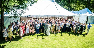 Wedding guests cheering with the bride and groom outside a large white marquee tent on a sunny lawn.