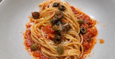 Authentic spaghetti alla puttanesca with tomato sauce, black olives, and capers in a white bowl.