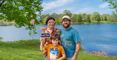 Staged photo of a couple with a young child in front of a lake, holding up paper props