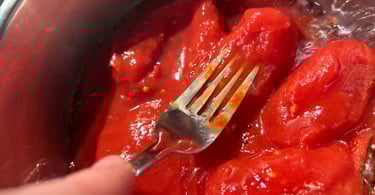 A metal fork mashing whole peeled San Marzano tomatoes in a stainless steel pot for a homemade pasta sauce.