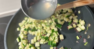 A metal ladle pouring broth over diced zucchini and onions cooking in a skillet.