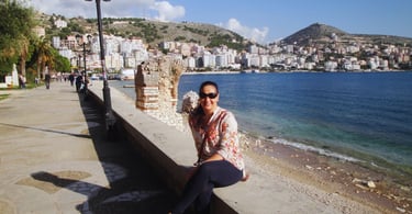 Autora sentada no píer de Sarandë, contemplando a paisagem tranquila da costa albanesa.
