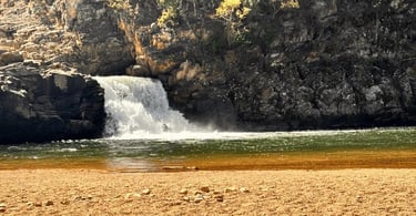 Cachoeira do Zé Carlinhos com queda d’água entre pedras escuras, poço verde e faixa de areia