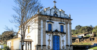 Pequena igreja histórica branca e azul ao lado de uma árvore seca em Lobo Leite, MG