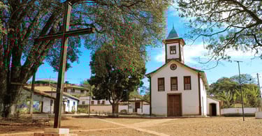 Capela com cores brancas e vermelhas, cruz de madeira a frente e arvores em uma praça em MG