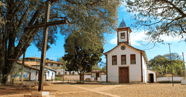 Capela com cores brancas e vermelhas, cruz de madeira a frente e arvores em uma praça em MG
