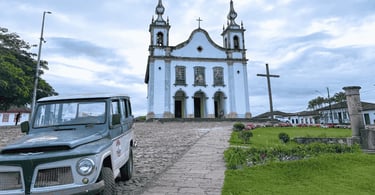 Igreja histórica em Catas Altas MG com cruz, jardim, calçamento de pedra e veículo antigo