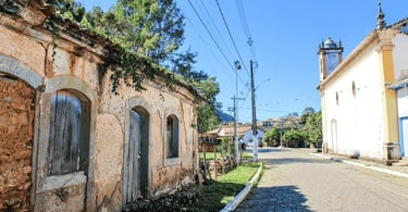 Rua de paralelepípedos em Itatiaia com uma casa histórica de pau-a-pique em primeiro plano e igreja