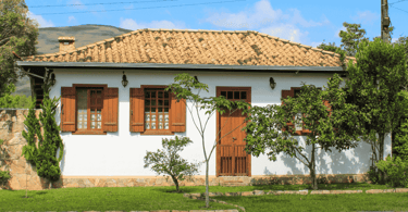 Casa branca, com vegetação em frente, janelas e porta de cor marrom, em Chapada MG