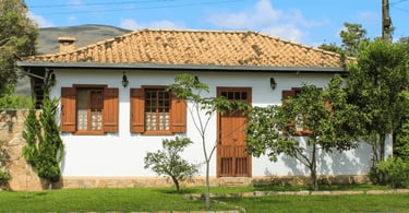 Casa branca, com vegetação em frente, janelas e porta de cor marrom, em Chapada MG