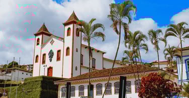 Igreja histórica em Serro com fachada branca, detalhes vermelhos, palmeiras altas e céu nublado