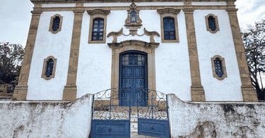 Fachada da Igreja Matriz de Glaura MG com porta e portão azul, detalhes em pedra e muro antigo
