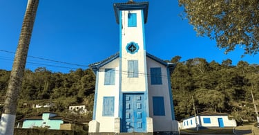 Igreja Matriz de Santo Antônio em Tapera, com cores brancas e azuis, vegetação ao fundo e casas