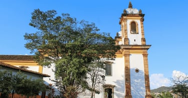 Igreja histórica em Sabará, com fachada branca, detalhes em amarelo, torre alta e árvore em frente