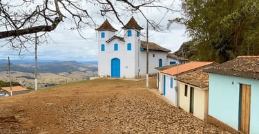 Igreja branca com portas azuis na Serra do Carola MG, cercada por casinhas simples e paisagem