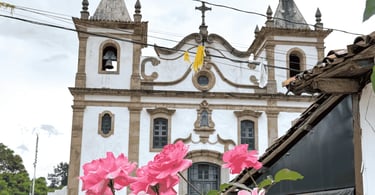 Igreja Matriz de Glaura MG com fachada colonial, torres com sinos e flores rosas em primeiro plano