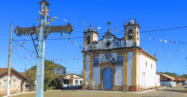 Igreja Matriz de Itatiaia com cores brancas e amarelas, com a cruz de cor azul a frente e céu aberto
