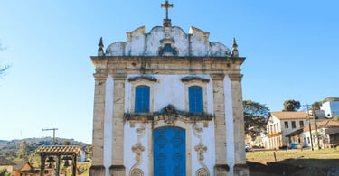Igreja histórica em Lobo Leite, MG, com fachada branca, porta azul e detalhes em pedra sob céu limpo