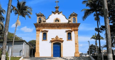 Igreja Matriz de Mateus Leme com fachada colonial branca, porta azul, sinos e palmeiras ao redor