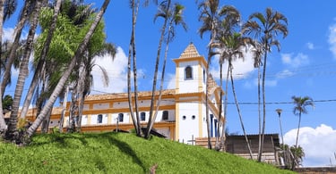 Igreja Matriz de Piedade do Paraopeba, com cores brancas e amarelas, coqueiros a frente