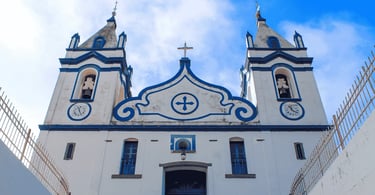 Fachada da Igreja Matriz branca com contornos em azul e duas torres sineiras. Raposos, MG