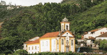 Igreja histórica em Ouro Preto MG com fachada branca e amarela, cercada por casas e morro verde