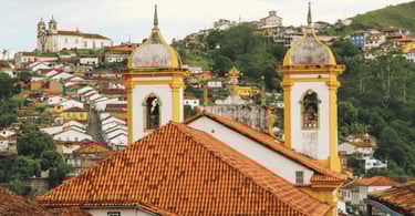 Igrejas históricas em Ouro Preto, com casas e rua de pedra ao lado direito, igreja branca e amarela