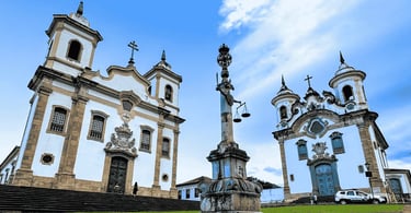 Igrejas históricas na Praça Minas Gerais em Mariana, com pelourinho ao centro e céu azul ao fundo