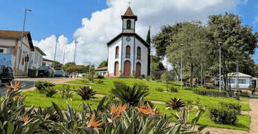 Igreja em Santa Bárbara vista da praça, com flores em primeiro plano, gramado e céu azul com nuvens