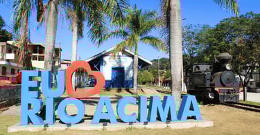 Locomotiva a vapor antiga estacionada ao lado de letreiro turístico de Rio Acima e serra ao fundo