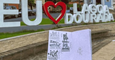 Passaporte da Estrada Real aberto, com o letreiro colorido da cidade de Lagoa Dourada ao fundo