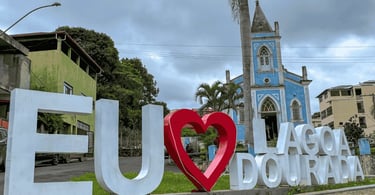 Placa escrita Eu Amo Lagoa Dourada, com igreja ao fundo, cidade nacional do rocambole em MG