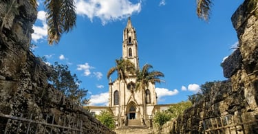 Fachada do Santuário do Caraça vista entre muros de pedra, com palmeiras, céu azul e nuvens brancas