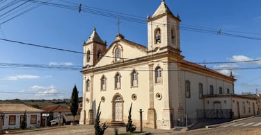 Vista lateral da Igreja Matriz de Nazareno, com duas torres, sinos e casarões históricos ao redor