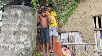  boys standing by cement water tank beside traditional West Bengal house.