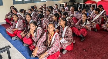 Around 30 girls in uniform sitting on floor in classroom.