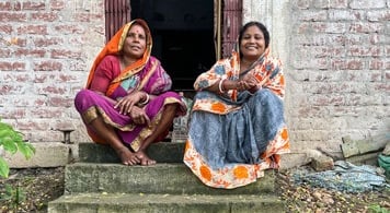 2 women in sarees sitting on step In front of building..
