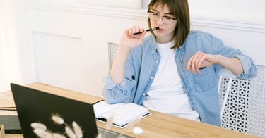 A focused woman wearing glasses and a denim shirt working at a wooden desk with a laptop and notebook.