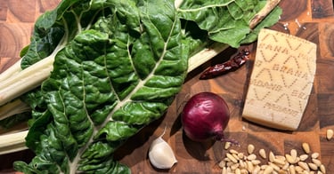 Fresh green chard leaves with a red onion, garlic, pine nuts, and parmesan cheese on a wooden board.