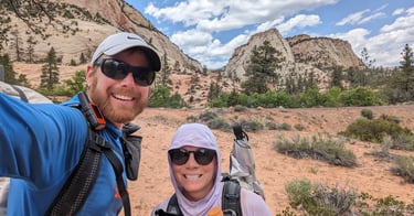 Moose and Bugs smiling in a selfie in front of beige sandstone features in Zion National Park.