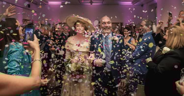 Smiling bride and groom celebrate with guests throwing colorful confetti at an indoor wedding reception.