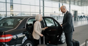 Professional chauffeur in a suit helping a senior woman into a luxury black sedan at the airport terminal.