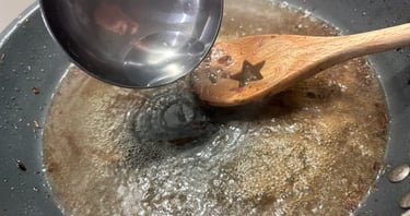 A metal ladle pouring water into a bubbling pan of broth for deglazing with a wooden spoon.