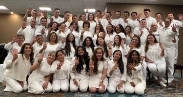 A large group of people in white clothing posing together and holding up four fingers for a photo.