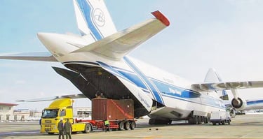 A Volga-Dnepr Antonov An-124 cargo plane loading a shipping container from a yellow truck at an airport.