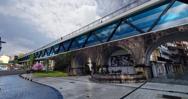Modern glass pedestrian bridge spanning over a historic stone archway in a European city plaza.